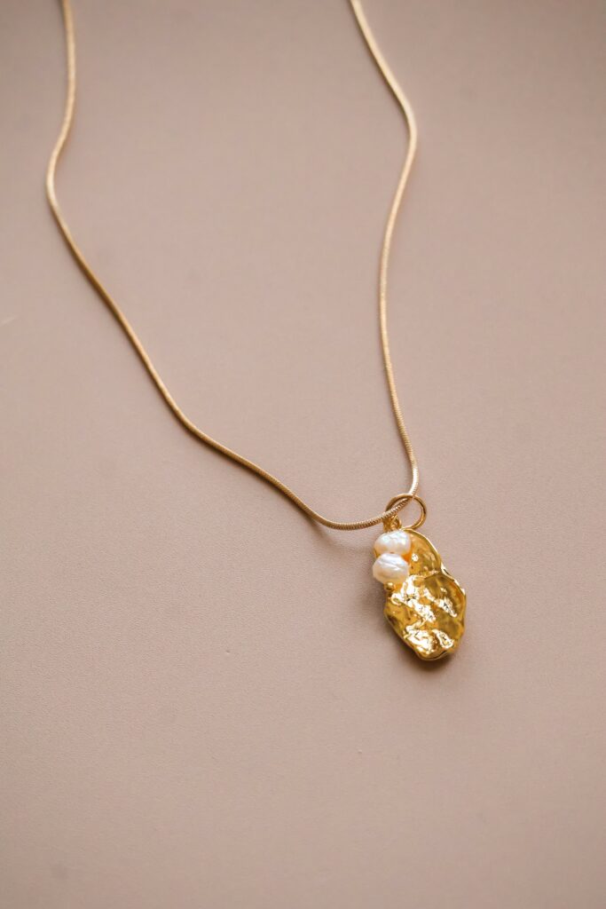 Gold necklace with a textured gold pendant and small white pearls on a beige background, close-up.
